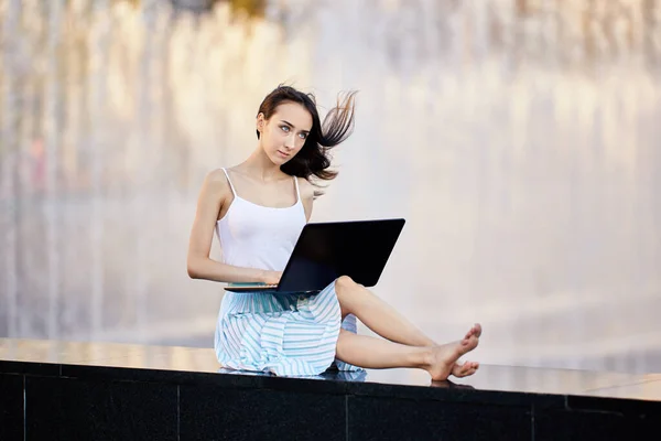 Schlanke Frau mit Laptop sitzt im Sommer am Brunnen im Stadtgarten. — Stockfoto