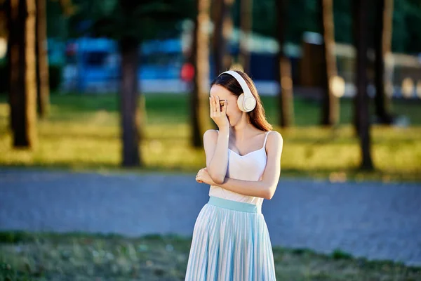 Śmiejąca się szczupła kobieta w słuchawkach bezprzewodowych stoi w parku. — Zdjęcie stockowe