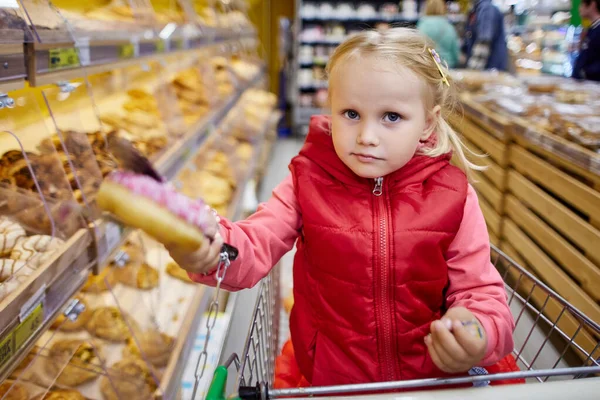 3 años de edad, niña en el carrito de la compra sostiene donute en el mercado. — Foto de Stock