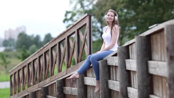 Donna con le cuffie penzola le gambe mentre si siede sulla recinzione nel parco pubblico. — Video Stock