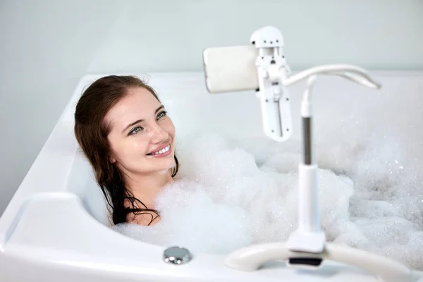 Felszerelés kéz ingyenes okostelefon tartó kád a fürdőszobában, fiatal nő vesz fürdés és videó megtekintése telefonon. — Stock Fotó