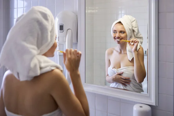 In Handtuch gehüllte Frau putzt sich vor Badezimmerspiegel die Zähne. — Stockfoto