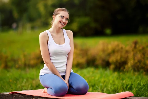 Mujer sonriente se sienta en la esterilla de ejercicio durante el yoga en el jardín de la ciudad. — Foto de Stock