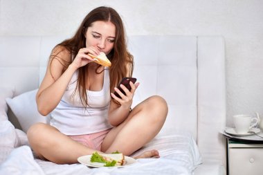 Akıllı telefonlu ve sandviçli aç bir kadın yatakta otururken kahvaltı yapıyor..