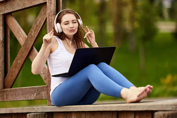 Vit kvinna med bärbar dator lyssnar på musik via trådlösa hörlurar i offentlig park. — Stockfoto