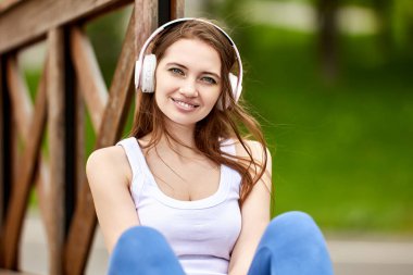 29 yaşında, kablosuz kulaklıklı gülümseyen bir kadın parkta oturuyor..