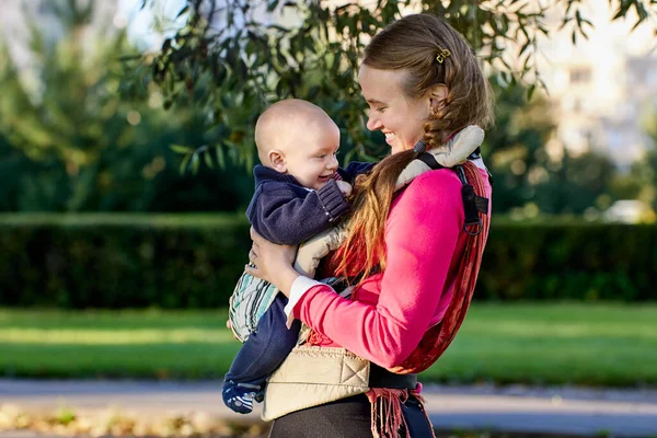 Child smiles in baby sling in hands of his mother during walking in park. — Stock Photo, Image