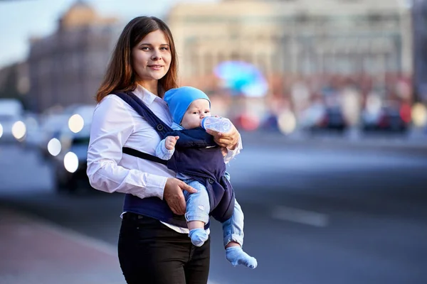 Lachende moeder is voeden baby in sling met behulp van fles in de buurt van het verkeer. Stockfoto