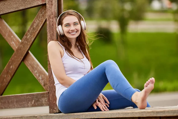 Uśmiechnięta biała kobieta w słuchawkach siedzi w parku latem. — Zdjęcie stockowe