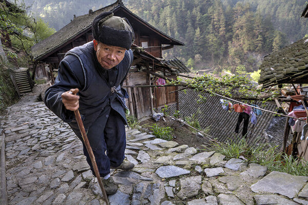 Aged Asian walks along stone road, laying on his stick.