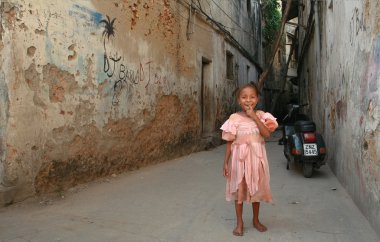 Afrikalı kız gülümsüyor, ayakta bir avlu içinde harap taş evler.