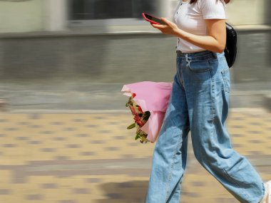 Bir buket çiçek ve cep telefonuyla yürüyen bir kadın. 