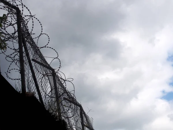 Yüksek bir duvarın tepesinde siluete sarılmış dikenli teller ve kara bulutlu bir gökyüzüne karşı çitler.