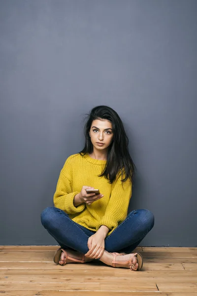 Calma chica de pelo negro con las piernas cruzadas y la célula mirando a la cámara —  Fotos de Stock