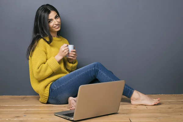 Feliz chica de pelo negro con taza en el suelo cerca de la computadora portátil —  Fotos de Stock