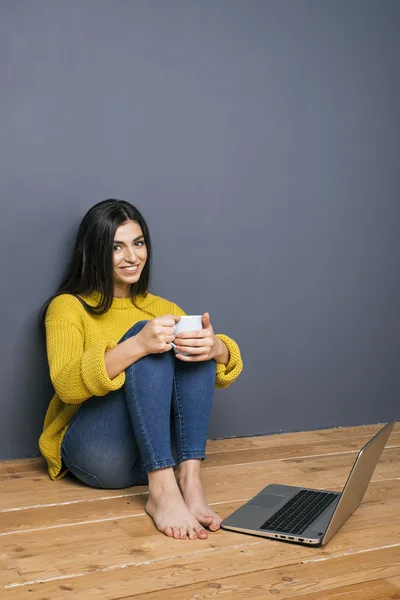 Barfota brunett med cup på golvet nära laptop — Stockfoto