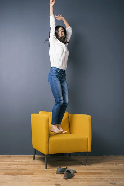 Glückliches Mädchen mit erhobenen Armen springt auf gelben Stuhl — Stockfoto