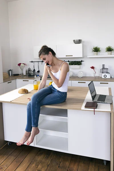 Mujer con las piernas cruzadas en la mesa de la cocina hablando por teléfono —  Fotos de Stock
