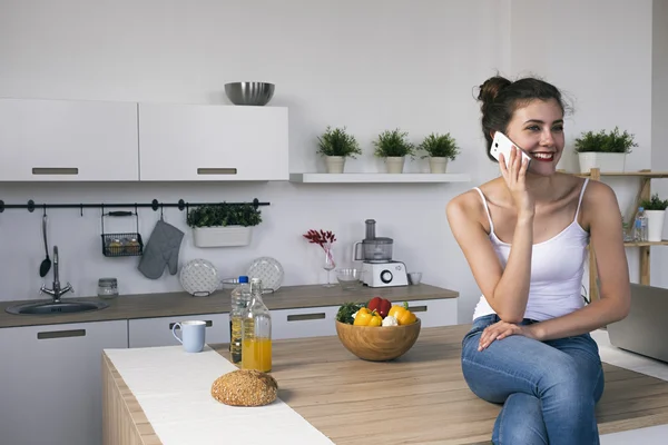 Sonriente morena hablando por la celda en la mesa de la cocina —  Fotos de Stock