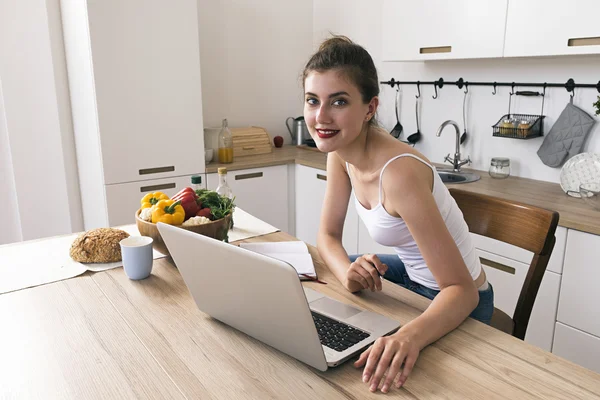 Vidám háziasszony mosolyogva kamera konyha asztal, laptop, élelmiszer és ital — Stock Fotó