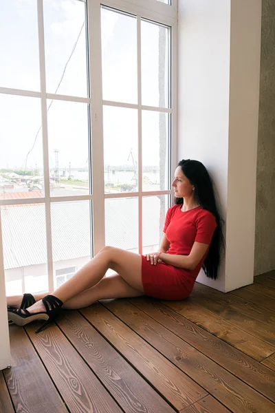 Vista lateral de la mujer elegante en vestido rojo en el suelo por la ventana — Foto de Stock