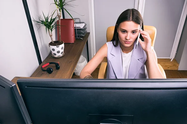 Upptagen brunett talar över cell och använda datorn i office — Stockfoto