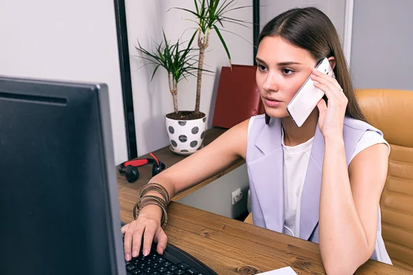 Schöne junge Frau telefoniert, während sie Computer benutzt — Stockfoto