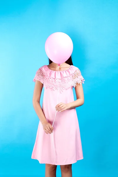 Conceptual shot of unrecognizable brunette in pink dress with pink balloon hiding her face on blue background.Isolate