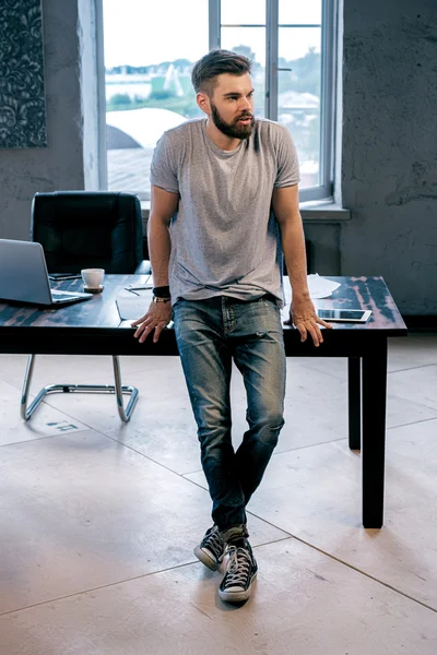 T恤和牛仔裤的英俊胡须男子的肖像靠在台式机上 笔记本电脑和平板电脑将目光移开 — 图库照片