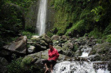 Ormanın ortasında şelalenin tazeliğinin tadını çıkaran kırmızı ceketli Asyalı bir adamın portresi. Bir gezgin büyük bir kaya nehrinde dinlenip sıcak kahveyi yudumlar.