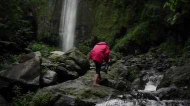Asyalı adam sıcak bir içecekle nehirdeki büyük kayaların arasında yürüyor. Kırmızı ceket giyen maceracılar ormanın ortasında bir şelalenin güzelliğinin tadını çıkarırlar.