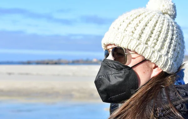 ブラックコート サングラス 白いウールの帽子で冬のビーチでCovid 19黒い顔のマスクを持つ女性 Kn95又はN95若しくはFfp2マスク ロイヤリティフリーのストック画像