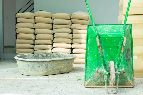 Green wheelbarrow and mortar mixing tank Pile of Cement in brown bags in a construction site with gray wall background