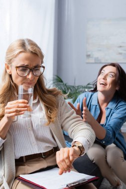 blonde woman therapist drinking water and checking time while brunette woman telling story with obsession clipart