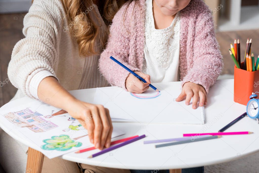 cropped view of little girl drawing pictures with colorful pencils while visiting psychologist