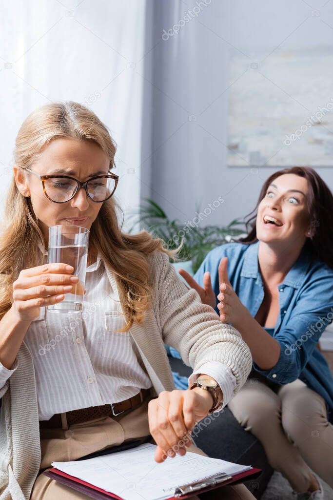 blonde woman therapist drinking water and checking time while brunette woman telling story with obsession