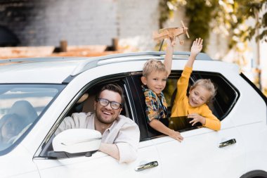 Smiling man looking at camera near wife and kids with toy in car  clipart