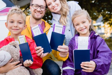 Smiling kids holding passports with air tickets near parents and car on blurred background outdoors  clipart
