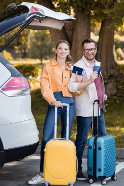 Çantalı ve pasaportlu gülümseyen bir çift açık hava arabasının yakınındaki kameraya bakıyor. 