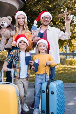 Noel Baba şapkalı neşeli bir aile ellerinde pasaport, valiz ve açık havada araba tutuyor. 