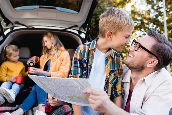 Smiling man holding map near son and wife with daughter near car on blurred background