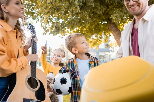 带着足球和柔软玩具的快乐孩子们站在父母身边 提着手提箱和带着音响的吉他 前景暗淡 — 图库照片