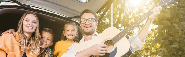 Homem Sorridente Tocando Guitarra Acústica Perto Família Porta Malas Carro — Fotografia de Stock