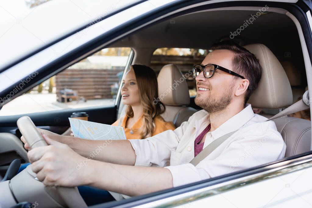 Smiling man driving car near wife with map and coffee to go on blurred background 