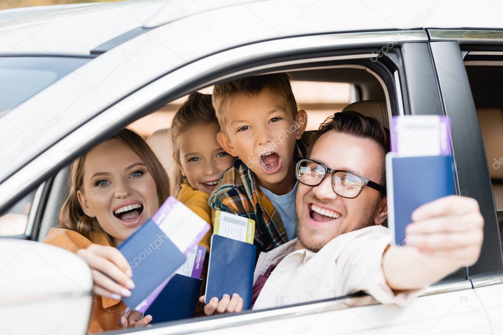 Cheerful family with kids holding passports with air tickets on blurred foreground in car 