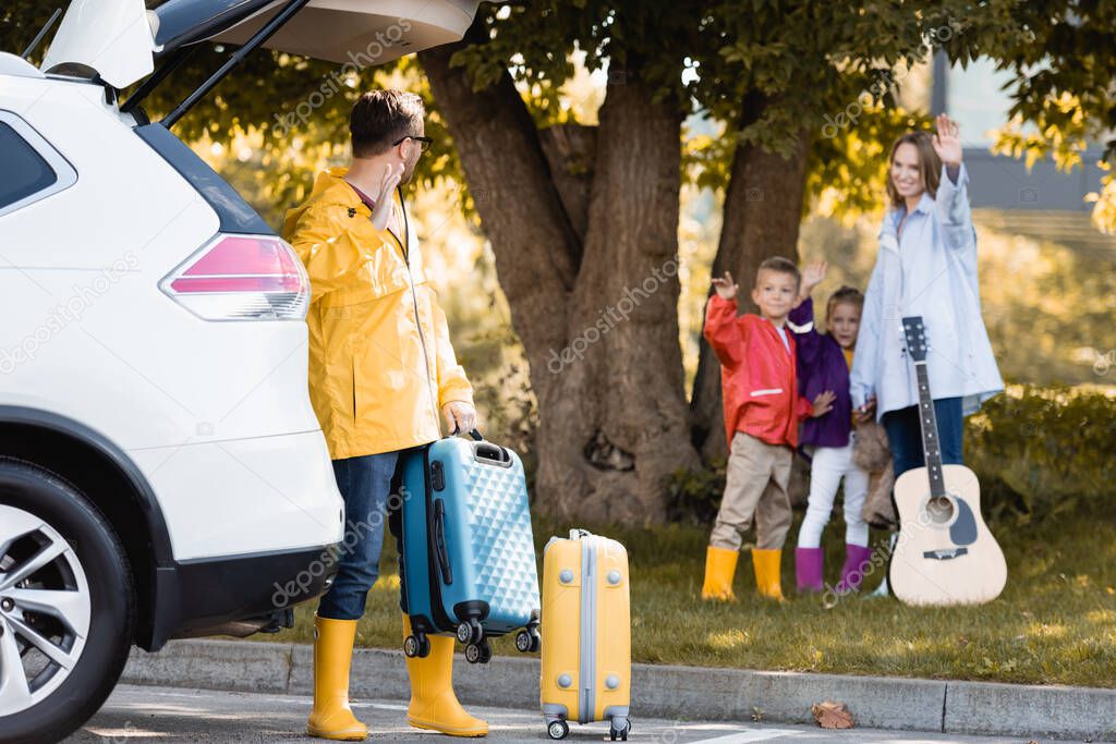 Man in autumn outfit waving hand while holding suitcase near car and wife with kids outdoors 