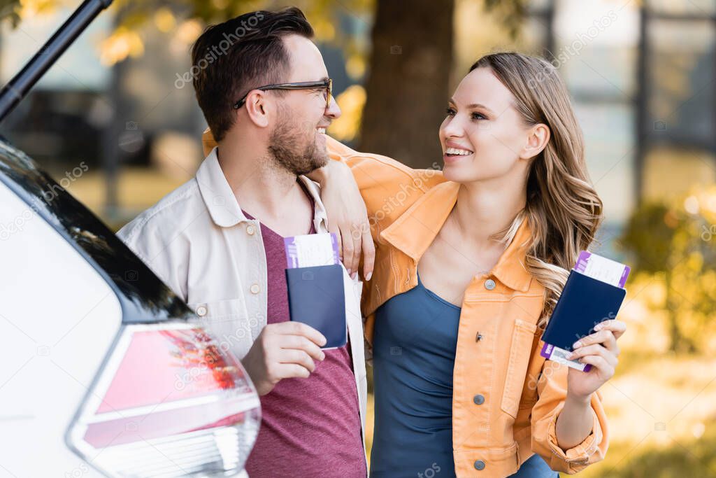 Smiling couple with passports and air tickets looking at each other near auto on blurred foreground outdoors 