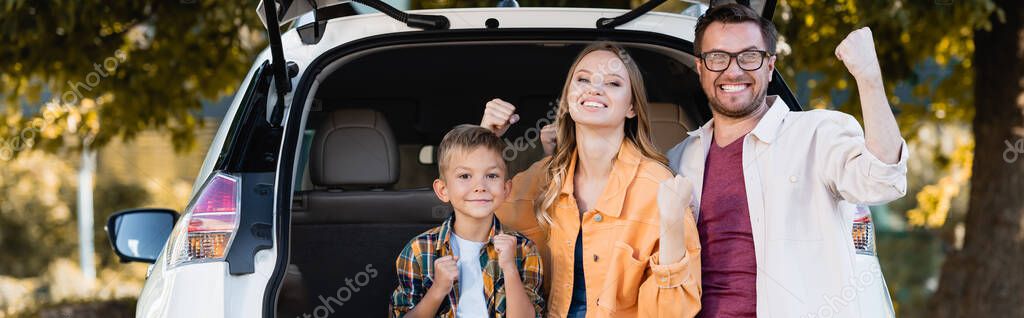 Smiling parents and soon showing yeah gesture near car outdoors, banner