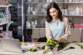 young florist making bouquet with eustoma flowers near laptop and racks on blurred background