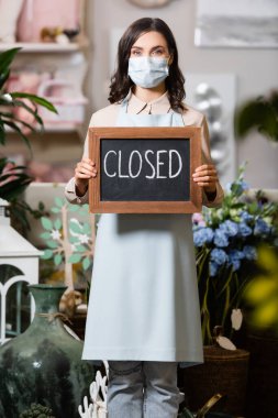 young florist in medical mask holding board with closed lettering in flower shop on blurred foreground clipart